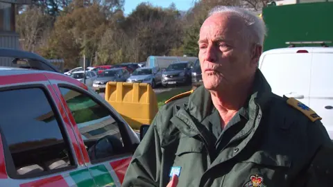 A man with white hair  wearing dark green ambulance attire standing in a car park.