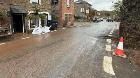 A road coloured brown in parts, with buildings to the left and a traffic cone on the right.