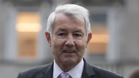 PA Media Michael Lowry pictured from the shoulders up, wearing a grey suit, pale shirt and a pink tie. He has short, grey hair. The background of the image is blurred. 