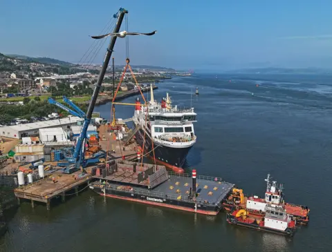 Douggie Choull A metal structure is lifted onto a barge by a large blue crane. A large ferry can be seen in the background, tied up at the quay. 