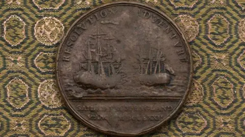 Auction House in Royal Wootton Bassett A medal with images of the ships used by James Cook on his second voyage. The medal is pleased on a green and yellow patterned cloth.