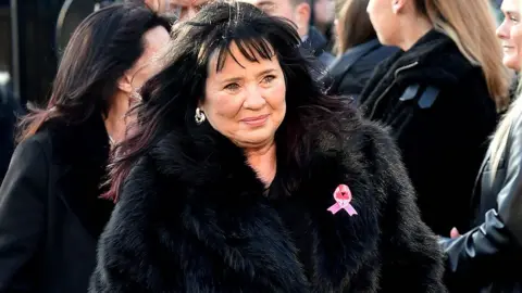 PA Media Coleen Nolan arrives at the church, wearing a black furry coat with a pink bow. She has long dark hair and is surrounded by mourners