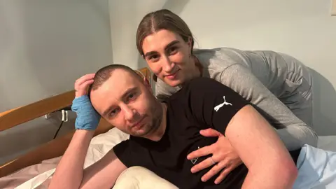 Man in black t-shirt lies on hospital bed with a woman in grey top crouching behind him holding his chest