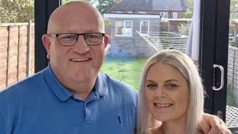 Heather Daly A couple stand in a kitchen with a bifold door and garden behind them. The man is wearing a blue polo neck top, is bald, and has wire framed glasses. She has white blonde hair shoulder length hair in a centre parting. A whirligig and shed can be seen in the garden beyond, and wooden fences either side.