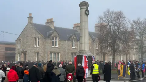 People gather around the war memorial in Grantown-on-Spey. Some of them are carrying poppy wreaths and flags. The memorial has flags draped around it at the bottom and a green wreath hanging near the top. There are houses in the background.