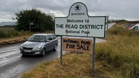 Jessica Peach A road sign which reads SKODA WELCOME TO THE PEAQ DISTRICT NATIONAL PARK. A cardboard sign is hung around the larger sign which reads NATIONAL PARK FOR SALE CASH NEEDED.