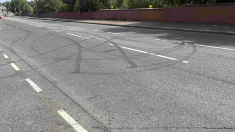 A road. On the road there are a number of dark tyre marks. Some of the tyre marks are in large circles.