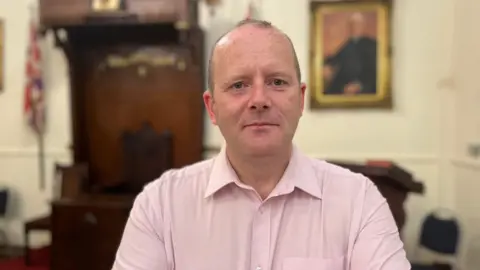 A balding man with grey hair, wearing a pink shirt sat in an office - the background is blurred but a framed image, wooden furniture and union flag are visible