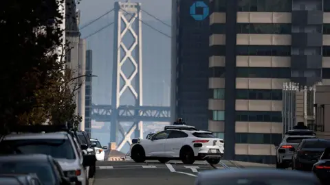 A white autonomous robotaxi driving on a city street with the Golden Gate Bridge and San Francisco Bay in the background. There are cars parked on the side of the street. 