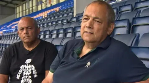 Dave and Pete sat side by side on navy blue seating inside a football stadium. Dave, on the left, is wearing a black tshirt with a Prostate Cancer UK logo - a white outline of a male silhouette - and Pete, on the right, is wearing a navy blue polo shirt with a Prostate Cancer UK badge. 