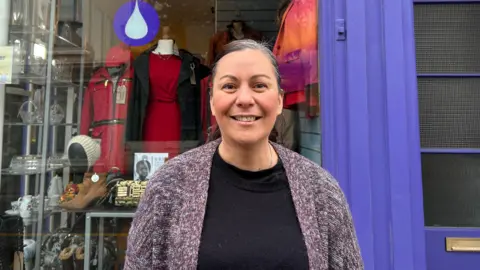 Elk Hargreaves is wearing a black top and purple-grey cardigan. She is stood in front of a charity shop with a purple facade.