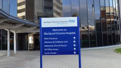 The exterior of Blackpool Victoria Hospital has a large round glass-fronted entrance beside a covered walkway and a blue sign saying 'Welcome to Blackpool Victoria Hospital'.
