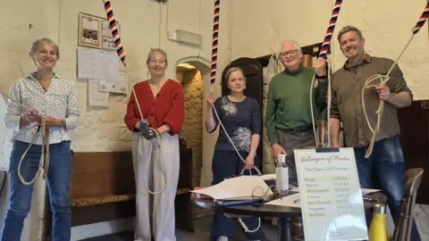 Joe Wheller Five people standing in a church holding on to ropes used for ringing bells. They are all smiling at the camera.