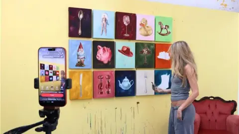 Patch Studio A woman with long blonde hair, wearing a grey vest and trousers, stands in front of a yellow wall, in front of a red armchair, with 15 oil paintings on the wall