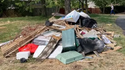 A pile of waste sits on a green space under a tree. The pile contains wooden struts and pallets, plastic boxes and bin bags among other items