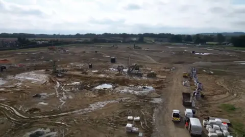 An aerial view of the One Horton Heath development. Shows a large section of land, which is mostly brown with rows of vans and goodsvehicles ot the right and crates piled with building materials. In the distance the land is lined with trees and greenery.