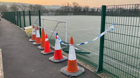 BBC A section of the green fence around the rugby pitch is flattened. Orange traffic cones are lined in front of the gap while blue and white police tape cordons off the scene.