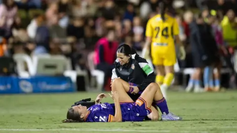 Mark Francesca Photographer Megan Wynne is lying on the ground with her leg held up by a physiotherapist. She is wearing purple football kit with orange stripes on her sleeve.