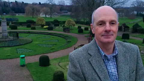 Wicksteed Park Oliver Wicksteed with short grey hair, wearing a grey jacket over a white and blue check shirt.  He is standing in front of a maze-like garden with paths around small dome hedges and trees.