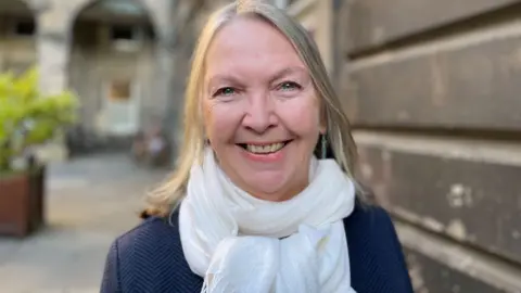 Jane Meagher is smiling at the camera. You can see her head and shoulders. She is wearing a blue suit jacket and white scarf. She has blonde shoulder-length hair.