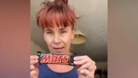 Victoria Gordon A woman holding up the old Mars Bar. She is wearing a navy vest and has red hair tied up with a full fringe.