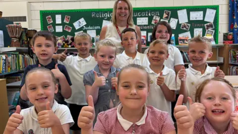 Lots of smiley school children huddle together with their thumbs up. A proud teacher stands behind them