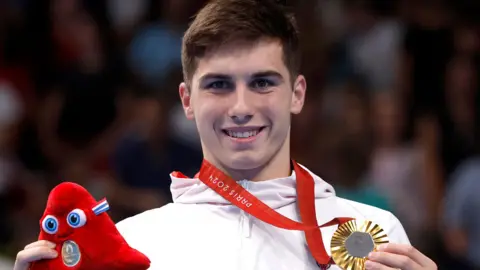 Will Ellard pictured in his white ParalympicsGB kit collecting his gold medal, following his 200m freestyle win at the Paralympics. He has short black hair and is smiling at the camera, clutching his medal in one hand and a toy Paralympics mascot in the other.
