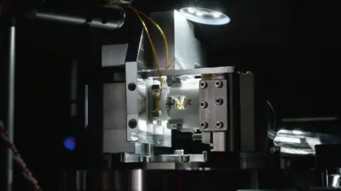 Wavelength Studio / Nu Quantum A qubit-photon interface. A piece of metallic hardware, lit by a single lamp in an otherwise dark room. In the centre of the image is a small metal and gold component, mounted inside white plastic, from which two wires emerge. The plastic is encased in a brushed metal surround. A lamp drops into the top of the image, shining light from the centre of the top of frame.