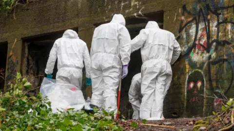 GMP Four crime scene investigators search Kersal Dale Bunker. Three are stood looking down at the ground while one is crouched down. The walls of the bunker are covered in graffiti.