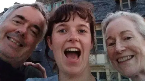 Burke Newman family A close-up smiling selfie of a young woman alongside her grinning parents. All three are pictured from the neck up and are standing in front of Mont-Saint-Michel, a tidal island and mainland commune in Normandy, France. 