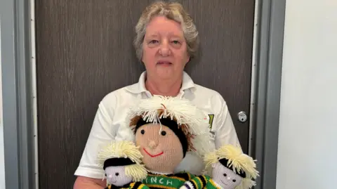 Carroll Weston/BBC Christine Carr, wearing a white top, with short fair hair, and holding three knitted version of Henry Pollock. She is standing by a door, that is closed. 