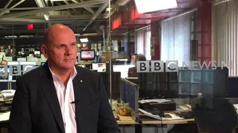 a man in a shirt and blazer looking at somebody offscreen. TV screens and the BBC News NI logo are visible behind him