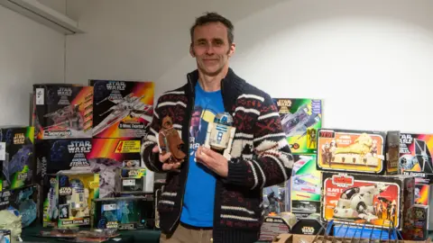 Jason Darling, a tall man with dark hair and sideburns. He has a blue star wars t-shirt on. He is holding action figures including R2-D2, a white robot character. Behind him are tens of boxes of action figures and vehicles from the series.