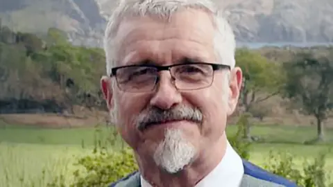 Close-up portrait of a person with short grey hair and glasses, wearing a suit and tie, standing outdoors with a blurred natural landscape of fields, trees, and hills in the background.