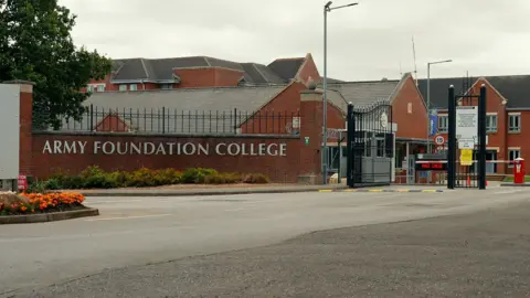 The words Army Foundation College are on a brick wall at the entrance to the campus. Large black gates are open with a red and white barrier slightly beyond. The college's buildings are behind.