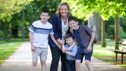 British Heart Foundation Mary McFarland with her three sons aged six, 10 and 12. All four family members are holding hands and smiling in a park on a sunny day. Mary is wearing a blue cardigan over a white top and dark trousers. The boys all have short, dark hair. The oldest two are wearing shorts and t-shirts while the youngest boy is wearing a blue shirt and trousers.