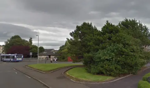 A footpath with a large tree in the middle. A road and bus are visible in the background