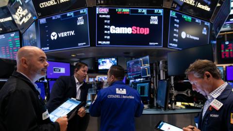 New York Stock Exchange traders in front of GameStop logo on screen.