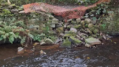 Bradford Council A stream with a retaining wall next to it that has partially collapsed and has been secured by orange netting