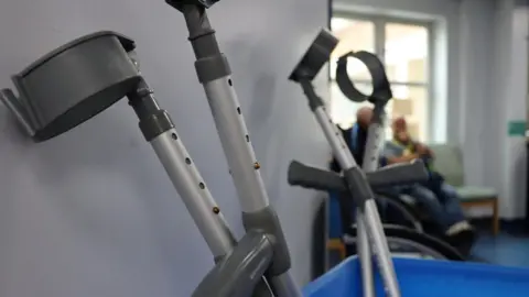 The picture shows to pairs of adjustable grey crutches with dark grey handles. They are leant against the wall and crossed over. 