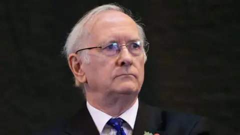 PA Media A man with white hair and glasses is wearing a black suit jacket, a white shirt and blue patterned tie