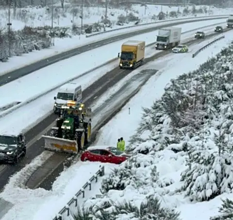 Fubar News A tractor beside a red car stuck in snow on a dual carriageway with other lorries in the background