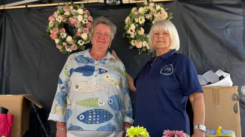 North East Lincolnshire Council Two women stood in a black tent smiling at the camera. The woman on the left has short, grey hair and is wearing a blouse with large blue fish on. The woman on the right had a short, blonde bob with a fringe. She is wearing a blue polo T-shirt and has her hand on the other woman's shoulder. Flower wreaths hang behind them. 