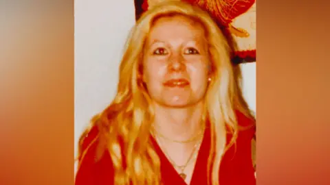 An old, heavily saturated photograph from the early 1990s of a woman with long blonde hair, who is wearing a stack of gold necklaces and a v-neck red top. She is looking above the shot and is slightly smiling.