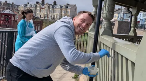 Mick English Matt Bailey, a man in his 30s, oiling the sage-green paint of a wooden fence with a little brush and a plastic pot of varnish. He is wearing bright blue surgical gloves.