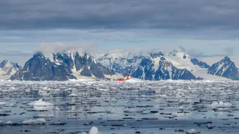 Elisenda Balleste A red research vessel is just visible in the distance, obscured by floating ice in flat-calm seas. The mountains of the Antarctic Peninsula, with huge flowing glaciers in between, as visible in the background