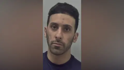 A police mugshot of a man wearing a dark blue top. He has black hair and is looking directly at the camera.