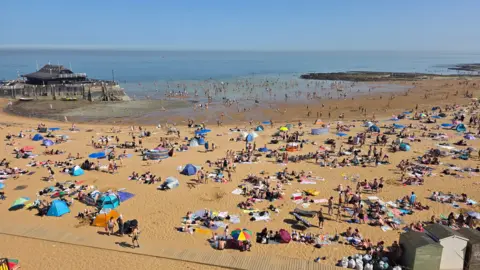 BBC Weather Watchers/mikea Viking Bay in Broadstairs on a sunny day. The beach is very busy, with hundreds of people sunbathing. The tide is out and the sky is blue.