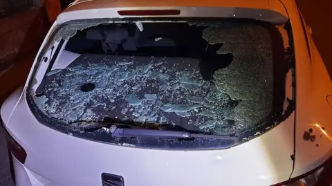 The picture shows a car window smashed. The car is a Seat and is white in colour.  The picture was taken at night time.