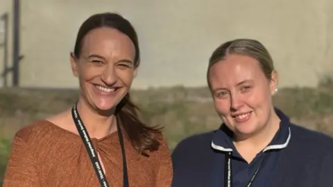 Shaunna Nicholson, Team Lead for the Integrated Wellness Service at Furness General Hospital, with Nakia Merchant, a Case Manager with the Integrated Wellness Service in Barrow. They are standing side-by-side and are both smiling at the camera. The background is blurred and the shot was taken outdoors in front of a patch of grass, with a building in the background. Ms Nicholson has shoulder-length dark hair in a ponytail and is wearing a burnt-orange dress. Ms Merchant is wearing a blue nurse uniform with a navy cardigan. Her dark-blond hair is tied back. They are both wearing lanyards around their necks.